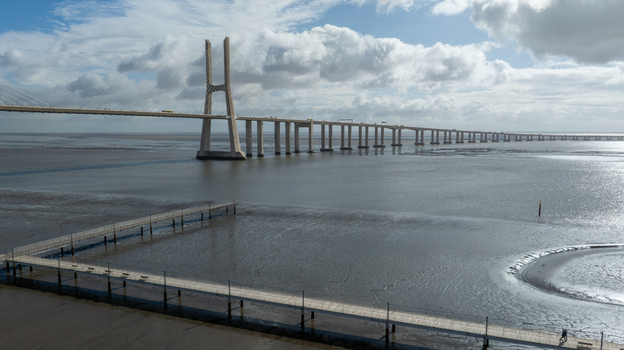 Aerial view of the Vasco da Gama Bridge spanning the Tagus River with a wooden walkway in the foreground under a cloudy sky in Lisboa Portugal.