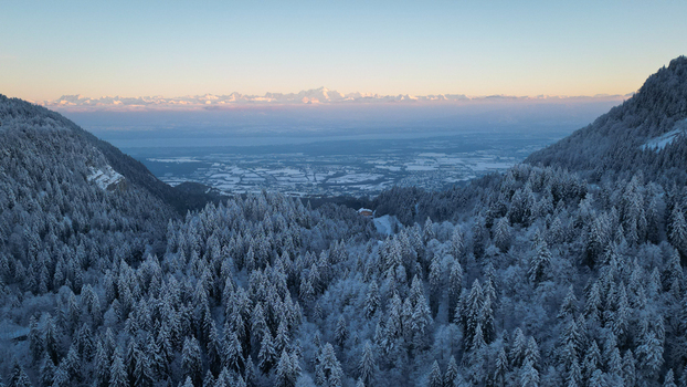 Aerial view of Col de la Faucille featuring snow covered pine forests and distant mountain peaks under a soft sunset sky in Gex Auvergne Rhone Alpes France. Aerial view of Col de la Faucille featuring snow covered pine forests and distant mountain peaks under a soft sunset sky in Gex Auvergne Rhone Alpes France.