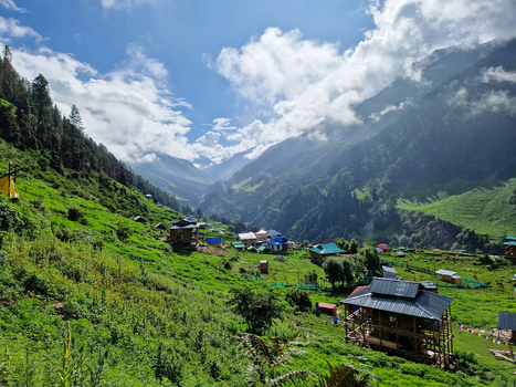 View of the historic village of Malana nestled in a lush green valley surrounded by towering Himalayan mountains under a blue sky with clouds Malana Himachal Pradesh India.