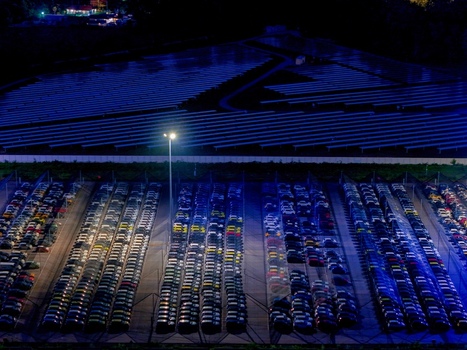 Aerial view of new cars parked at a distribution centre illuminated by bright floodlights at night with a solar panel field in the background in Rivalta di Torino Piedmont Italy.