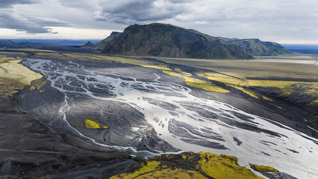 Aerial view of the braided river system at Thakgil featuring dark volcanic sand vibrant green moss and rugged mountains under a cloudy sky in Vik Myrdalshreppur Iceland.