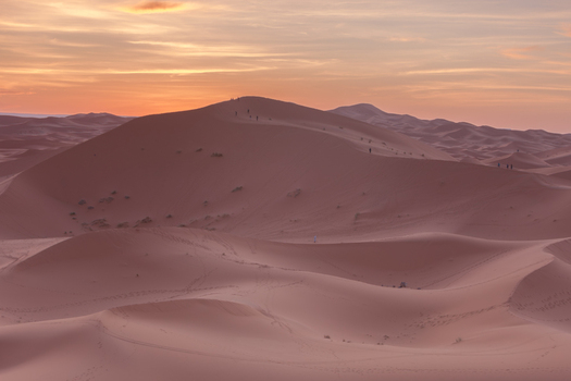 View of the Sahara desert dunes at sunset with people walking along the ridges under a warm golden sky in Morocco.