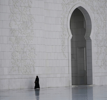 View of a woman in a black abaya walking past a white marble wall with intricate floral carvings and a large arched doorway in Abu Dhabi Abu Dhabi United Arab Emirates. View of a woman in a black abaya walking past a white marble wall with intricate floral carvings and a large arched doorway in Abu Dhabi Abu Dhabi United Arab Emirates.