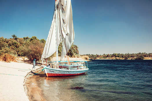 View of a traditional wooden felucca boat with a large white sail moored on the sandy riverbank of the Nile with people in Aswan Aswan Governorate Egypt. View of a traditional wooden felucca boat with a large white sail moored on the sandy riverbank of the Nile with people in Aswan Aswan Governorate Egypt.