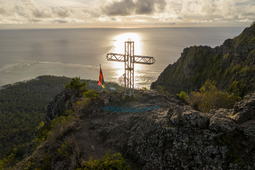 Aerial view of a metal cross and a Mauritian flag on a rocky mountain peak overlooking the Indian Ocean at sunset Moka District Mauritius. Aerial view of a metal cross and a Mauritian flag on a rocky mountain peak overlooking the Indian Ocean at sunset Moka District Mauritius.