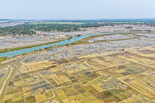 Aerial view of the intricate patchwork of agricultural fields meeting the winding river with wind turbines standing tall in the distance Cox s Bazar Chittagong Division Bangladesh. Aerial view of the intricate patchwork of agricultural fields meeting the winding river with wind turbines standing tall in the distance Cox s Bazar Chittagong Division Bangladesh.