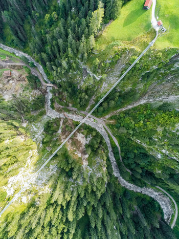 Aerial view of a long suspension bridge spanning a deep mountain valley with a river and dense pine forests in Austria. Aerial view of a long suspension bridge spanning a deep mountain valley with a river and dense pine forests in Austria.