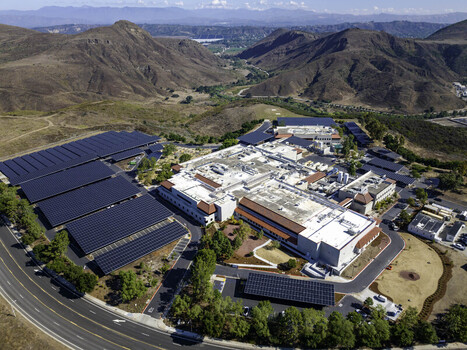 Aerial view of a pharmaceutical company facility featuring extensive solar panel arrays surrounded by arid hills and green trees in Los Angeles California United States. Aerial view of a pharmaceutical company facility featuring extensive solar panel arrays surrounded by arid hills and green trees in Los Angeles California United States.