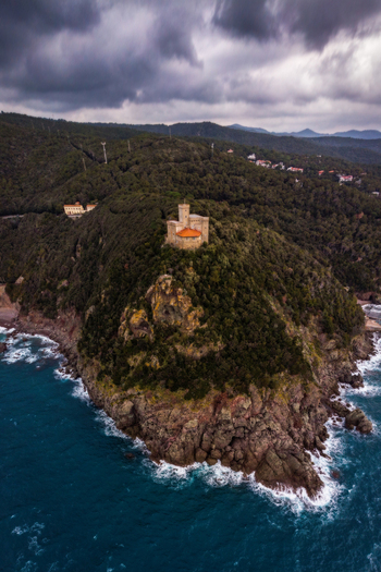 Aerial view of the historic Torre del Boccale perched on a rugged cliff overlooking the blue Mediterranean sea under a dramatic cloudy sky in Calafuria Toscana Italy.