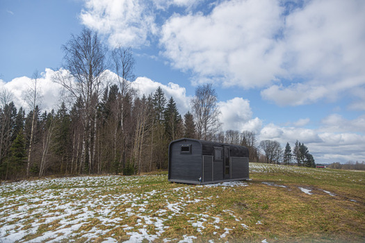 View of a modern dark colored tiny house cabin in a rural field with patches of snow and a forest background in Latvia. View of a modern dark colored tiny house cabin in a rural field with patches of snow and a forest background in Latvia.