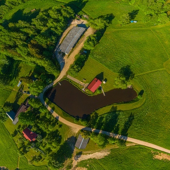 Aerial view of a rural landscape featuring a small pond red roofed houses lush green fields and surrounding trees under bright sunlight Latvia. Aerial view of a rural landscape featuring a small pond red roofed houses lush green fields and surrounding trees under bright sunlight Latvia.