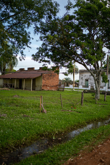 View of a weathered brick building with a rusted roof set in a grassy field with a drainage ditch and large trees in Guaviravi Corrientes Province Argentina.