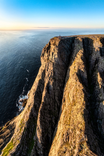 Aerial view of the rugged North Cape cliffs illuminated by golden hour light with deep rock fissures and the vast Arctic Ocean below in Nordkapp Municipality Finnmark Norway. Aerial view of the rugged North Cape cliffs illuminated by golden hour light with deep rock fissures and the vast Arctic Ocean below in Nordkapp Municipality Finnmark Norway.