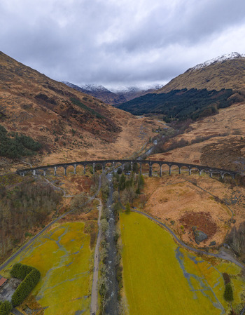 Aerial view of the curved Glenfinnan Viaduct spanning a valley with a river and rugged hills under a cloudy sky in Glenfinnan Scotland United Kingdom.