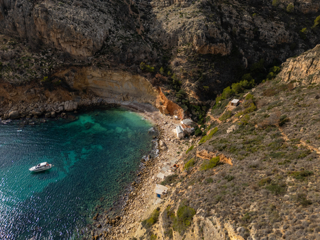 Aerial view of Cala Llebeig with turquoise waters rocky cliffs and small traditional houses along the pebble beach in El Poble Nou de Benitatxell Valencian Community Spain.