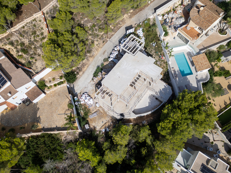 Aerial view of a residential construction site with a concrete structure and scaffolding surrounded by villas with swimming pools and pine trees in Moraira Valencian Community Spain.
