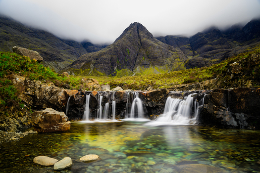 View of the crystal clear water of the Fairy Pools cascading over dark volcanic rocks with the misty Cuillin mountains in the background on the Isle of Skye Scotland. View of the crystal clear water of the Fairy Pools cascading over dark volcanic rocks with the misty Cuillin mountains in the background on the Isle of Skye Scotland.