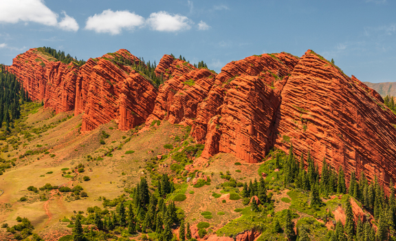 Aerial view of the red sandstone Seven Bulls Rock formation featuring dramatic jagged cliffs green slopes and pine trees under a clear blue sky in Jeti Oguz Issyk Kul Region Kyrgyzstan.