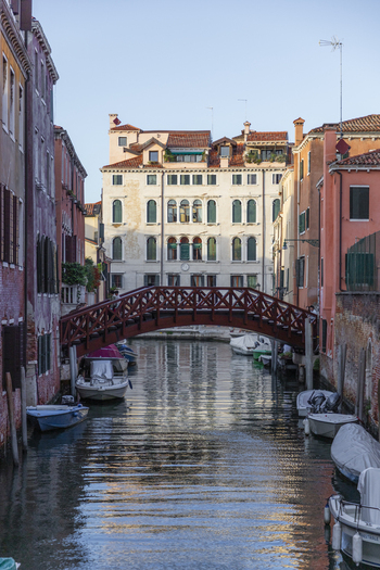 View of a wooden footbridge crossing a narrow canal in Venice lined with historic buildings and moored boats under a clear sky in Venice Veneto Italy. View of a wooden footbridge crossing a narrow canal in Venice lined with historic buildings and moored boats under a clear sky in Venice Veneto Italy.