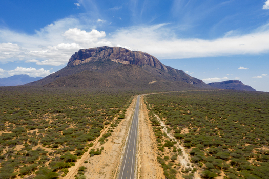 Aerial view of a straight road stretching through the arid scrubland towards the prominent volcanic mountain in Marsabit Marsabit County Kenya. Aerial view of a straight road stretching through the arid scrubland towards the prominent volcanic mountain in Marsabit Marsabit County Kenya.