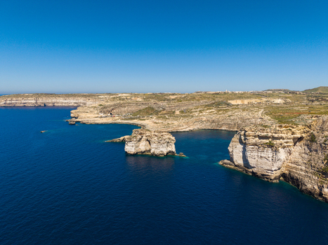 Aerial view of the rugged limestone cliffs and sea stack at Dwejra Bay overlooking the deep blue Mediterranean sea under a clear sky in Gozo Region Malta. Aerial view of the rugged limestone cliffs and sea stack at Dwejra Bay overlooking the deep blue Mediterranean sea under a clear sky in Gozo Region Malta.