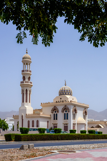View of a mosque with a tall minaret and ornate dome surrounded by manicured hedges under a clear blue sky in Muscat Muscat Governorate Oman. View of a mosque with a tall minaret and ornate dome surrounded by manicured hedges under a clear blue sky in Muscat Muscat Governorate Oman.