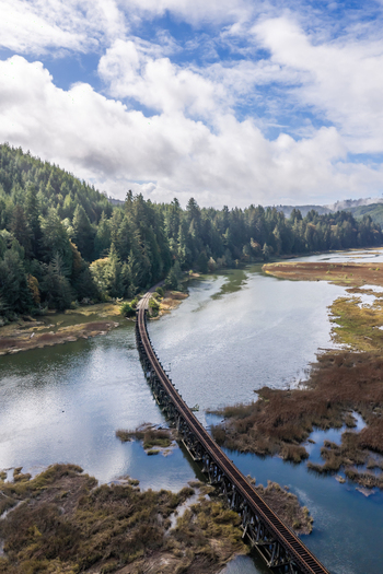 Aerial view of railroad tracks winding across wetlands near the Siuslaw River in Florence Oregon with calm water marshland and evergreen forest along the Oregon Coast.