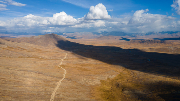 Aerial view of the vast Deosai Plains with a winding dirt road cutting through the golden landscape under a dramatic cloudy sky in Skardu Gilgit Pakistan.