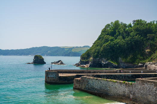 View of the historic stone harbor walls and calm turquoise waters with lush green cliffs and a rocky coastline in the background in Charlestown Cornwall United Kingdom. View of the historic stone harbor walls and calm turquoise waters with lush green cliffs and a rocky coastline in the background in Charlestown Cornwall United Kingdom.