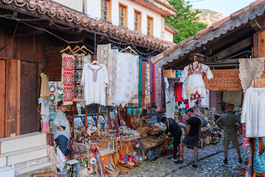 Kruje Albania 12 June 2026 View of the Old Bazaar with traditional textiles and souvenirs displayed outside wooden shops along a cobblestone street with people browsing.