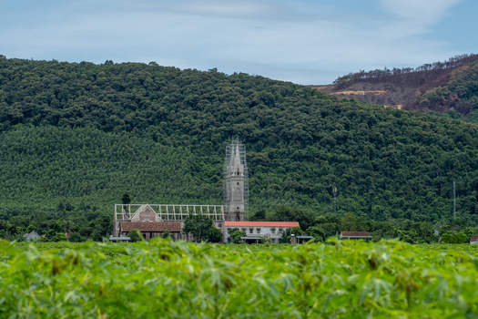 View of a church with a tall spire under renovation with scaffolding nestled against a backdrop of lush green forested hills in Vietnam. View of a church with a tall spire under renovation with scaffolding nestled against a backdrop of lush green forested hills in Vietnam.