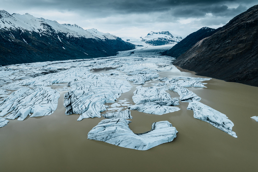 Aerial view of Vatnajokull glacier with icebergs floating in a glacial lake surrounded by rugged mountains under a dramatic cloudy sky in Iceland.