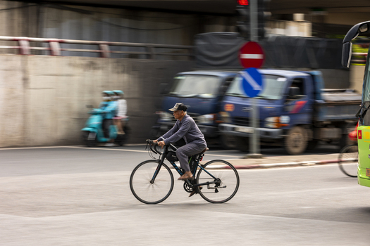 Hanoi Ha Noi Vietnam 11 November 2025 View of an elderly man riding a bicycle through a busy urban intersection with motion blurred traffic and trucks in the background.