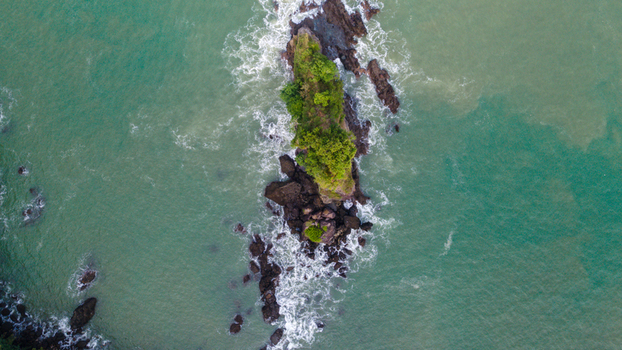 Aerial view of the rocky Karang Agung beach islet covered in lush green vegetation surrounded by turquoise sea water and white waves in Ayah Central Java Indonesia. Aerial view of the rocky Karang Agung beach islet covered in lush green vegetation surrounded by turquoise sea water and white waves in Ayah Central Java Indonesia.