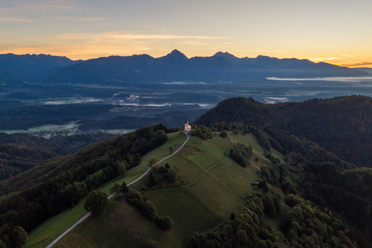 Aerial view of the Church of Saints Primus and Felician on a hilltop with a winding road surrounded by green slopes and forests under a golden hour sky in Jamnik Kranj Slovenia.
