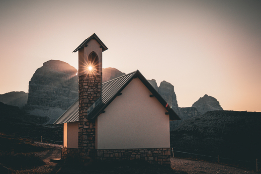 View of the small stone church at Tre Cime di Lavaredo with a sunburst shining through the bell tower against the dramatic mountain peaks in Auronzo Veneto Italy.
