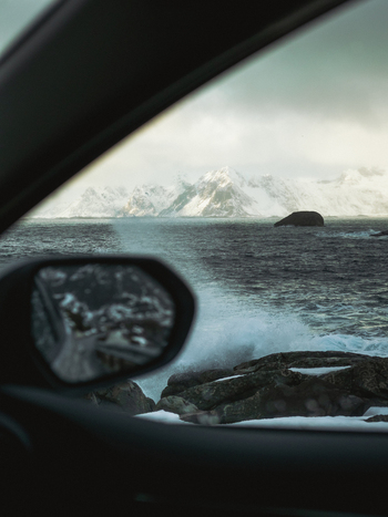 View of icy waves crashing on rocky shores framed by a car window against distant snow capped mountains Henningsv&aelig;r Nordland Norway.