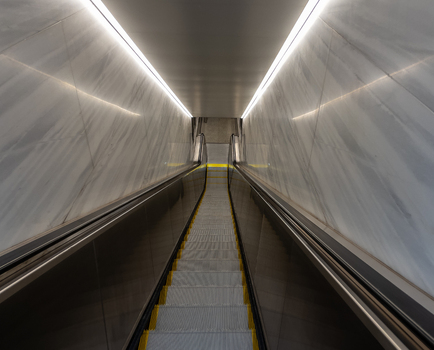 View of a modern subway escalator descending between polished marble walls with bright linear lighting in New York New York United States. View of a modern subway escalator descending between polished marble walls with bright linear lighting in New York New York United States.