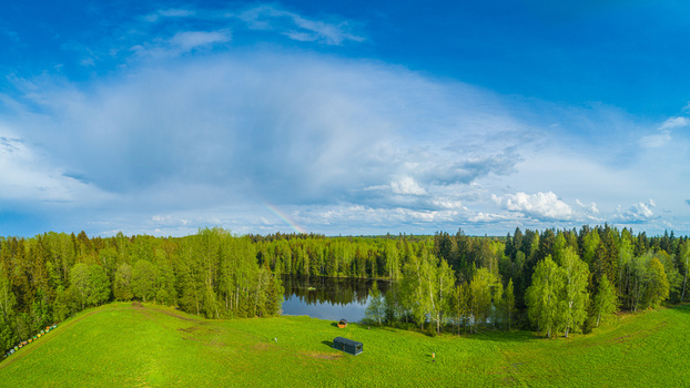 Aerial view of a lush green field with a small pond and dense forest under a bright blue sky with a faint rainbow in Latvia. Aerial view of a lush green field with a small pond and dense forest under a bright blue sky with a faint rainbow in Latvia.