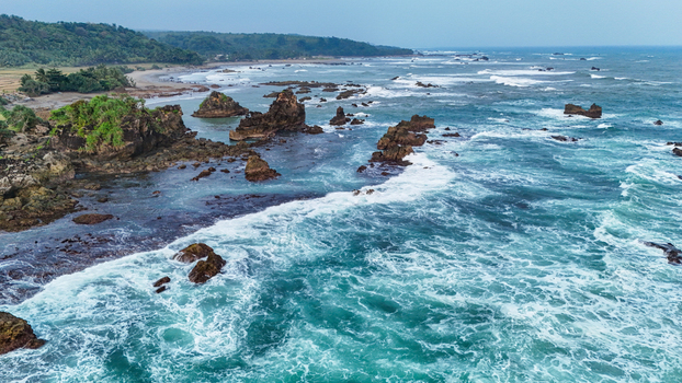 Aerial view of a rocky beach with turquoise ocean waves crashing against jagged rock formations along the lush tropical coastline of Indonesia. Aerial view of a rocky beach with turquoise ocean waves crashing against jagged rock formations along the lush tropical coastline of Indonesia.