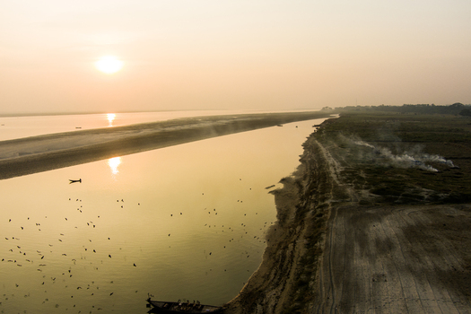 Aerial view of the Padma River at sunset with a small boat on the water birds flying and smoke rising from the riverbank in Rajshahi Rajshahi Division Bangladesh.