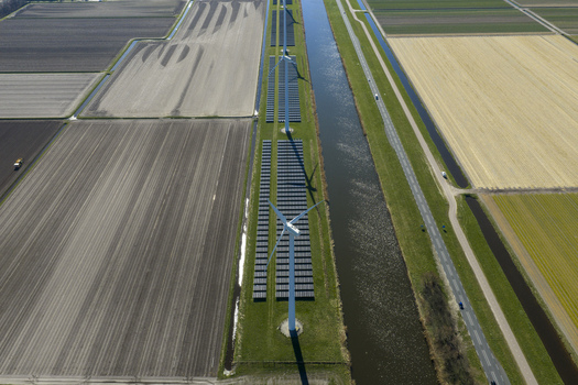 Aerial view of wind turbines and solar panel arrays situated along a canal amidst vast agricultural fields under bright sunlight in Netherlands. Aerial view of wind turbines and solar panel arrays situated along a canal amidst vast agricultural fields under bright sunlight in Netherlands.