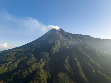 Aerial view of Mount Merapi volcano with smoke rising from the crater and lush green forested slopes under a clear blue sky Kecamatan Pakem Daerah Istimewa Yogya Indonesia. Aerial view of Mount Merapi volcano with smoke rising from the crater and lush green forested slopes under a clear blue sky Kecamatan Pakem Daerah Istimewa Yogya Indonesia.