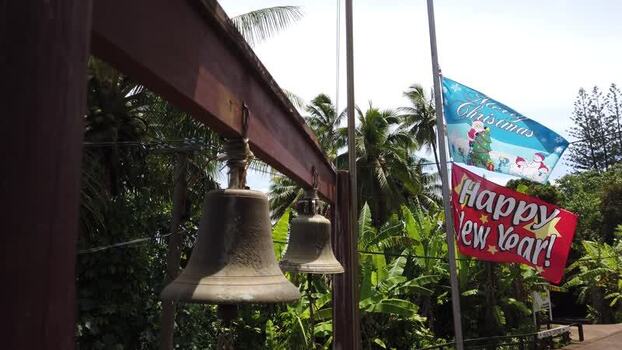 Aerial view of weathered bells hang amidst the lush tropical foliage with the vibrant Merry Christmas and Happy New Year flags waving gently Rarotonga Cook Islands.