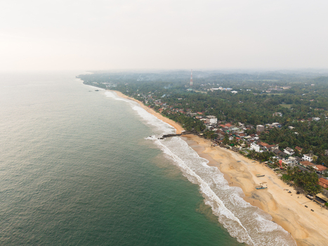 Aerial view of the tropical coastline with a sandy beach palm trees and coastal buildings under a hazy sky in Galle Southern Province Sri Lanka.