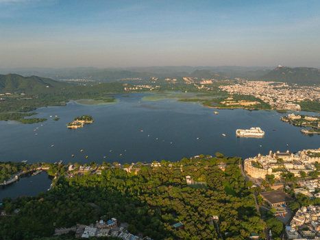 Aerial view of Lake Pichola with the historic City Palace and Lake Palace surrounded by lush greenery hills and the urban landscape of Udaipur Rajasthan India.