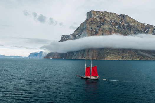 Aerial view of a sailboat with red sails navigating the deep blue waters of a fjord near the massive Storoen mountain with clouds clinging to rocky cliffs in Avannaata Kommunia Greenland. Aerial view of a sailboat with red sails navigating the deep blue waters of a fjord near the massive Storoen mountain with clouds clinging to rocky cliffs in Avannaata Kommunia Greenland.