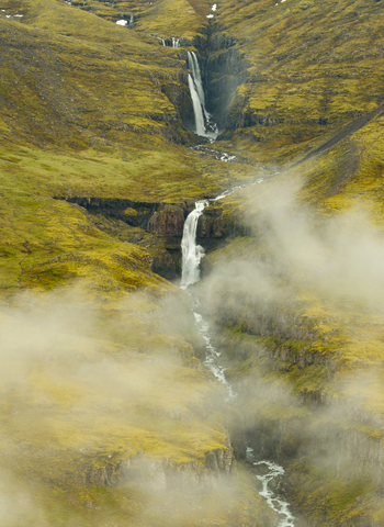 Aerial view of a cascading waterfall flowing through a lush green mossy mountain gorge in the Eastern Region of Iceland. Aerial view of a cascading waterfall flowing through a lush green mossy mountain gorge in the Eastern Region of Iceland.