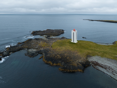 Aerial view of a stark white lighthouse with a red roof stands sentinel on a rugged green topped island against the steely grey sea K&aacute;lfshamarsv&iacute;k H&uacute;nabygg&eth; Iceland.
