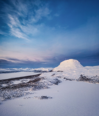 Aerial view of the snow covered Saana fell rising above the frozen landscape and lake under a vast blue sky in Kilpisjarvi Lapland Finland.